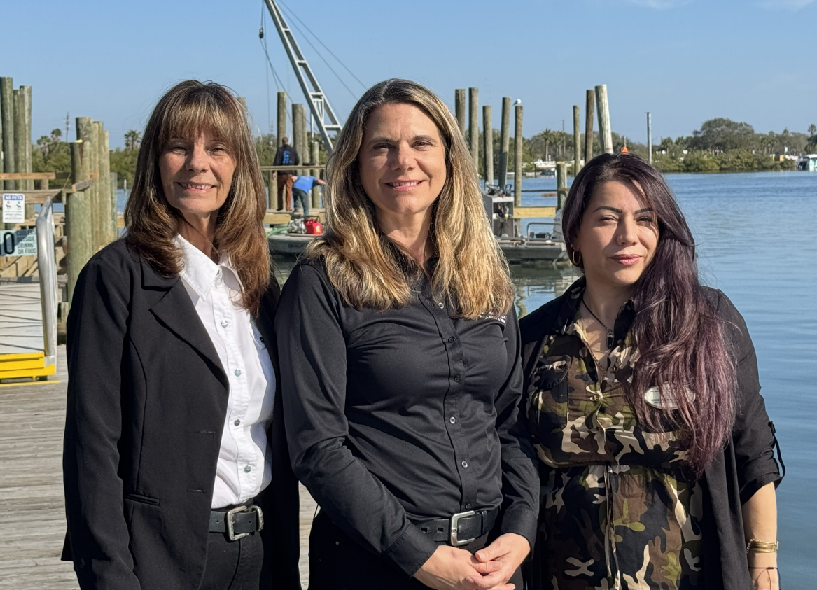 Three team members from the Riance Realty team stand together on a sunny marina dock in Florida, dressed in professional attire. The background features calm blue water, wooden pilings, and boats, reflecting their connection to Florida’s waterfront and residential real estate market.
