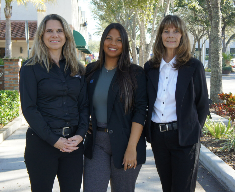 Three professional team members from the Riance Realty team standing outdoors on a sunny sidewalk lined with trees and shops. Dressed in smart business attire, they smile warmly, representing excellence and professionalism in Florida real estate.