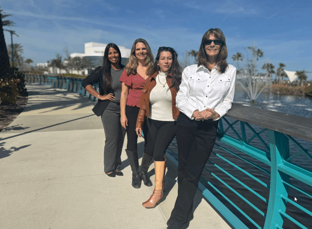 A group photo of four professional women from the Riance Realty team standing on a sunny waterfront walkway with palm trees and a fountain in the background. They are dressed in stylish business-casual attire, smiling confidently, representing real estate services in Florida.